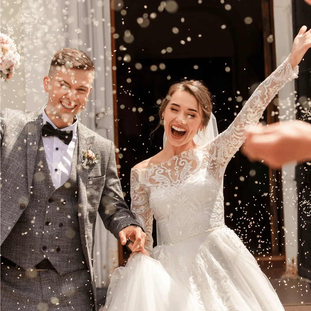 A smiling bride and groom holding hands as they walk out of a building under falling confetti.