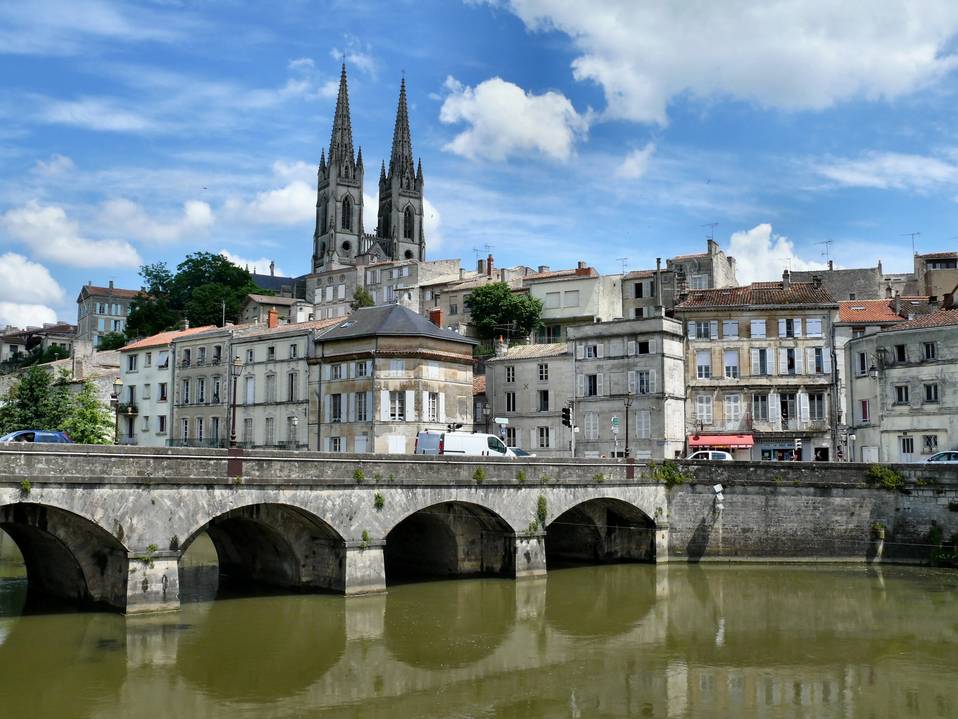 A stone bridge with arches crosses a river in front of historic buildings, with a large cathedral featuring twin spires rising in the background under a partly cloudy sky.