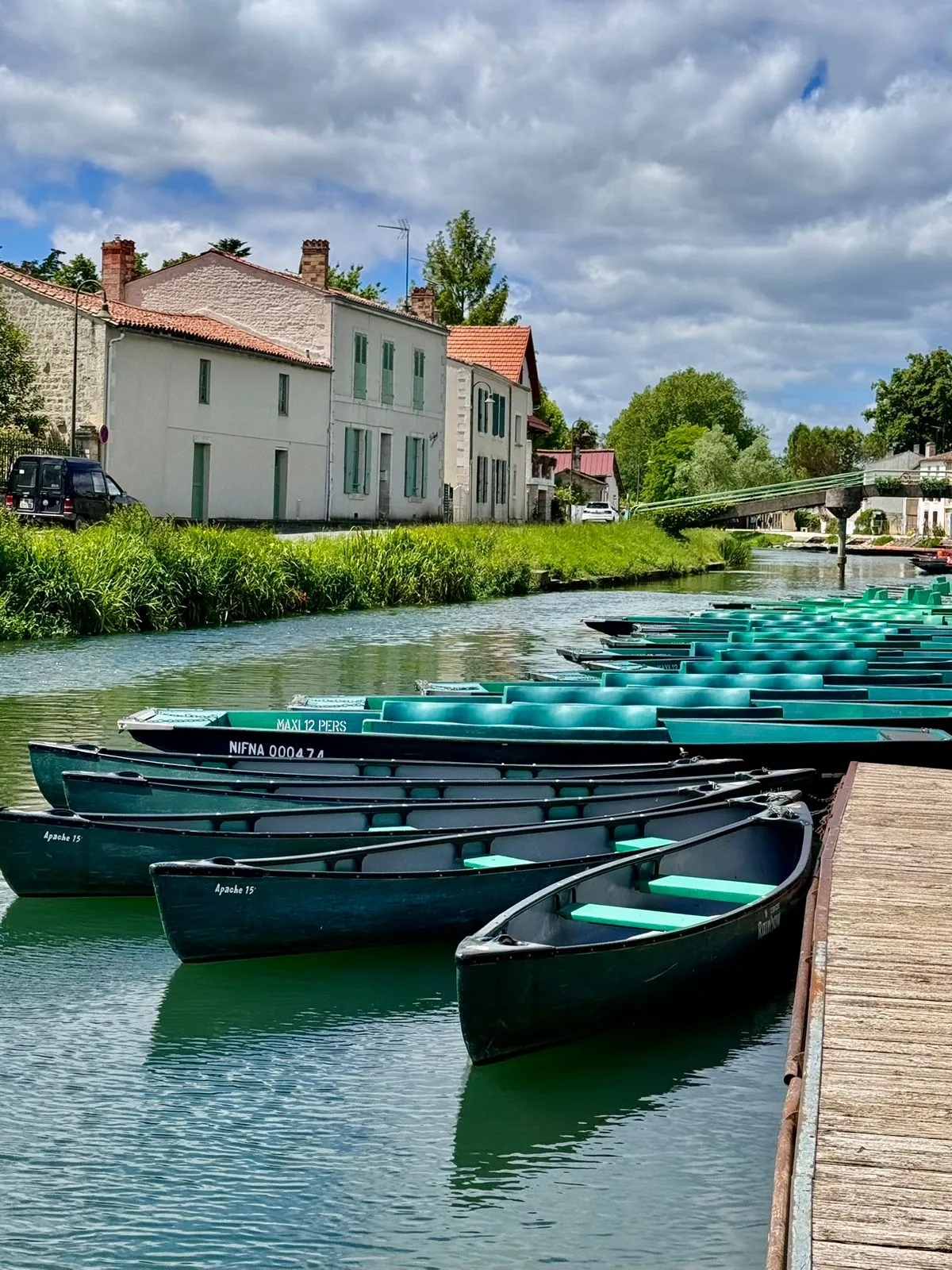 A row of green boats is moored along a wooden pier on a calm canal, with white houses and greenery lining the opposite bank under a partly cloudy sky.