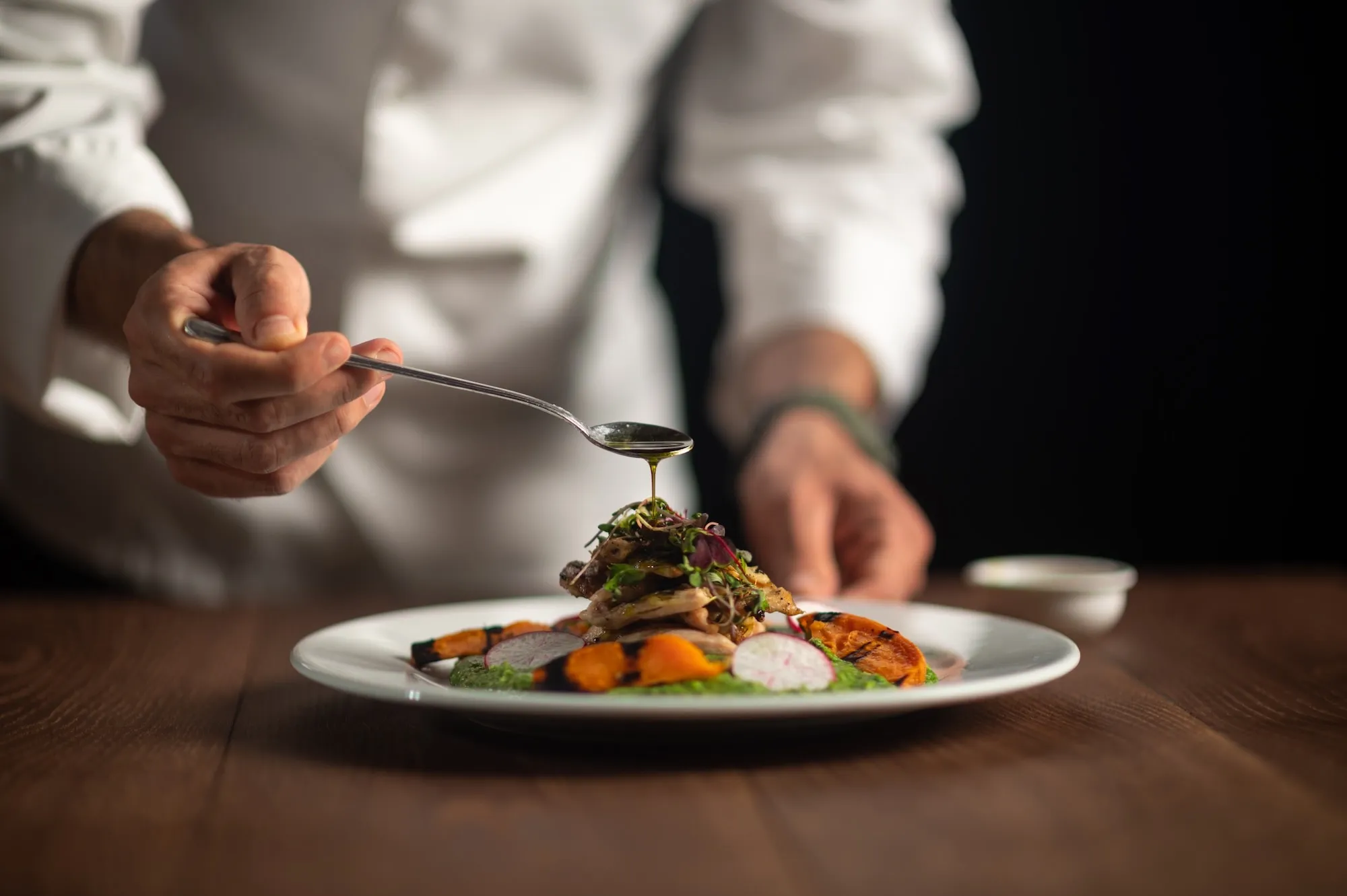 A chef in a white coat drizzles sauce over a plated gourmet dish with vegetables and greens on a white plate, set on a wooden table against a dark background.