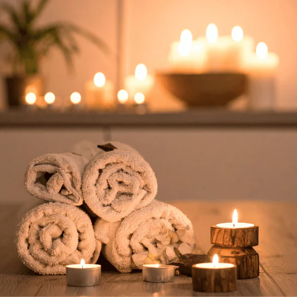 Rolled white towels arranged on a wooden surface with burning candles and tea lights in the background, creating a warm, relaxing spa ambience.