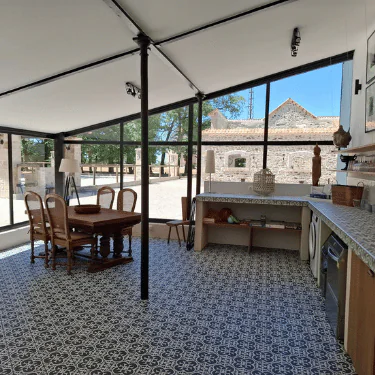 Modern kitchen and dining area with patterned tiled floor, wooden table and chairs, large windows, and outdoor view of stone buildings under blue sky. Worktop with shelves and kitchen appliances on the right.