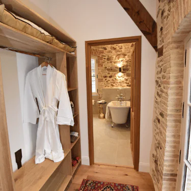 A wooden wardrobe with a white dressing gown hangs next to a doorway leading to a rustic bathroom with a freestanding bath, stone walls, and a wall-mounted light.