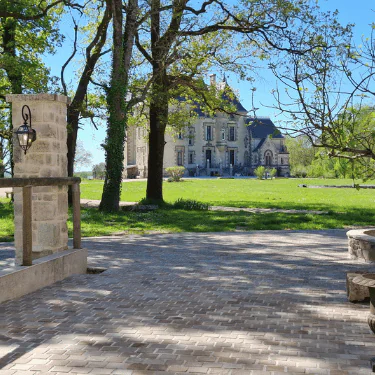 A stone courtyard with a lantern and well overlooks a large manor house surrounded by trees and green lawn under a clear blue sky.