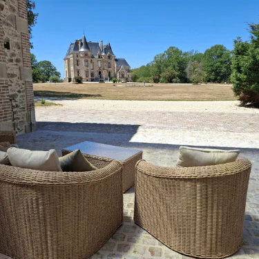 Two wicker chairs and a table on a stone terrace overlook a large lawn with an historic château in the background under a clear blue sky.