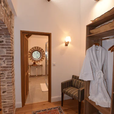 A cosy dressing area with a striped chair, bathrobe, and open wardrobe. An exposed brick wall and doorway lead to a bright bathroom with a round window and basin visible.