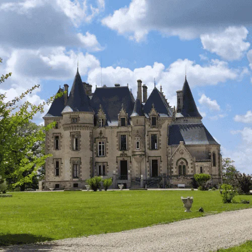 A stone château with multiple turrets and steep roofs stands on green grass under a partly cloudy blue sky, with a gravel path and some trees in the foreground.
