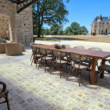 A long wooden outdoor dining table with chairs on a stone patio, shaded by a canopy. There’s outdoor seating nearby, large trees, and a grand building visible in the background.