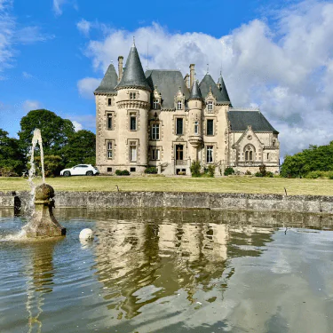 A historic stone château with turrets stands behind a round fountain, reflecting in the water. A white car is parked on the grass, and trees surround the scene under a partly cloudy blue sky.