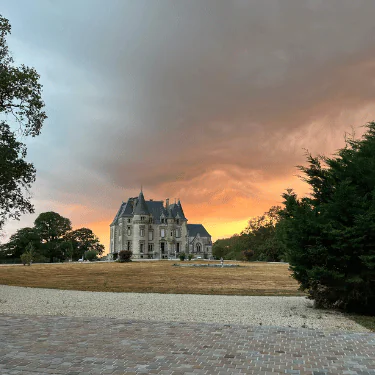 A grand stone château with pointed towers sits on a grassy lawn, framed by trees. The sky glows orange and pink at sunset, with dramatic clouds overhead and a stone path in the foreground.