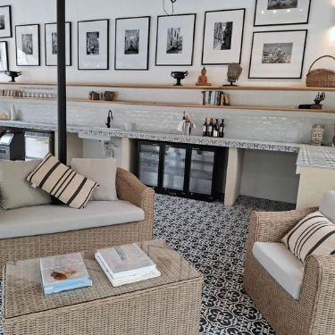 Cosy indoor seating area with wicker furniture, patterned floor tiles, a glass coffee table with books, a long worktop, framed photos on the wall, and decorative items on shelves.