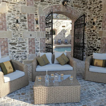 Wicker garden furniture with beige cushions and green pillows sits on stone flooring in front of large arched gates. A small swimming pool, sun loungers, and more stone walls are visible in the background.