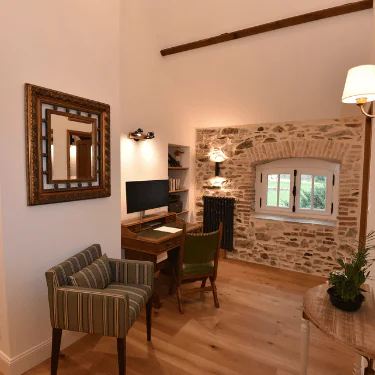 A cosy home office with a wooden desk, chair, and striped armchair. There's a computer monitor, bookshelves, and a window set in a stone wall, with wooden flooring and a decorative mirror on the white wall.