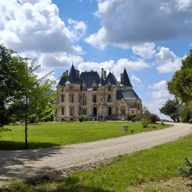 A stone château with pointed roofs sits on a large green lawn, surrounded by trees, under a blue sky with scattered clouds. A gravel drive curves in front of the building.