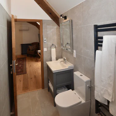 Modern bathroom with beige tiles, a grey vanity basin, a toilet, a wall mirror, towel rail with white towels, and an open door leading to a cosy room with wooden flooring and an armchair.