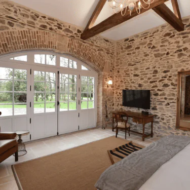 A cosy bedroom with stone walls, wooden beams, a desk with a TV, and large arched windows overlooking a green garden. A bed, armchair, and luggage rack complete the rustic decor.