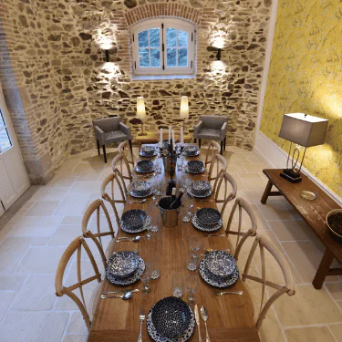 A long wooden dining table laid for eight with patterned crockery, in a rustic room with stone walls, a yellow feature wall, chairs, and soft lighting.