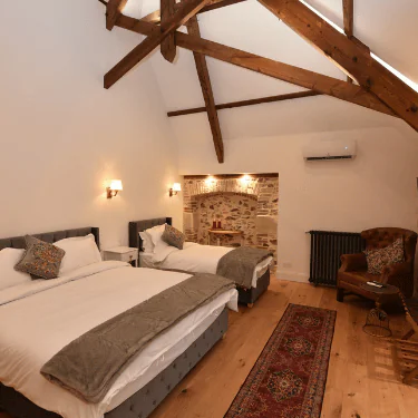 A cosy bedroom with two beds, exposed wooden beams, a brick feature wall, an armchair, a rug, and warm lighting. The room has wooden flooring and modern, rustic decor.