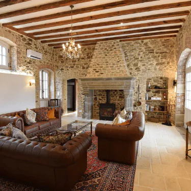 A rustic living room with stone walls, exposed wooden ceiling beams, leather settees, a large fireplace, chandelier, arched windows, and a Persian rug on a light stone floor.