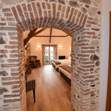 View through a brick and stone archway into a cosy bedroom with wooden floors, a bed, armchairs, and double glass doors opening to a garden outside. Exposed wooden beams decorate the ceiling.