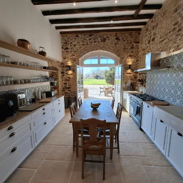 Rustic kitchen with stone walls, wooden beams, open shelves, white cupboards, a wooden table with chairs, modern appliances, and large windows overlooking a sunny outdoor scene.