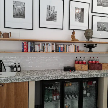 A worktop with a mini fridge containing drinks, books on shelves, framed black-and-white photos on the wall, a small sink, and decorative statuettes and bottles are visible in a modern kitchen space.