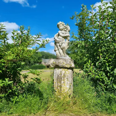 A weathered stone statue of two hugging children stands on a mossy plinth, surrounded by tall grass and green bushes, under a bright blue sky with scattered clouds.