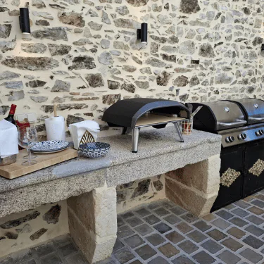 Outdoor kitchen area with a stone worktop, pizza oven, grill, plates, glasses, and utensils set up against a rustic stone wall on a cobbled patio.