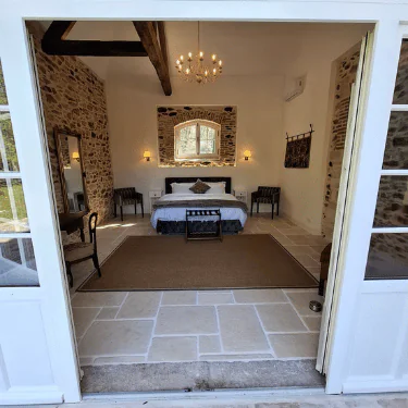A cosy bedroom with stone walls, a large bed, a chandelier, chairs, and a mirror, viewed through open double doors. A window above the bed lets in natural light.