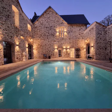 A large stone building with exterior lights reflecting in a rectangular swimming pool at dusk, creating a serene and inviting atmosphere.