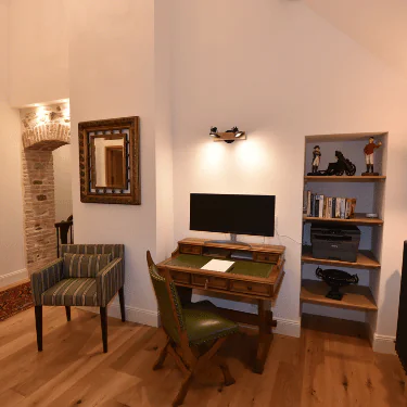 A cosy home office with a wooden desk, green chair, computer monitor, striped armchair, wall mirror, shelves with books, and decorative items. Warm lighting creates a welcoming atmosphere.