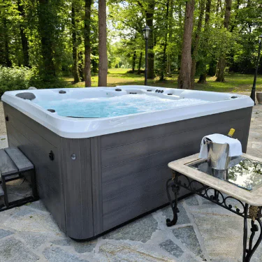 A bubbling hot tub sits on a stone patio in a wooded area, with a small table nearby holding a silver ice bucket and glasses.