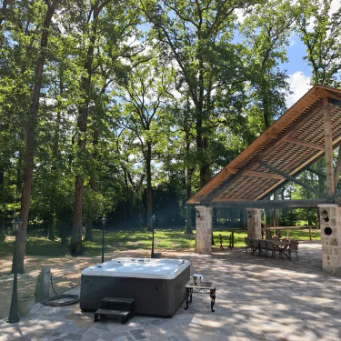 A hot tub sits on a stone patio near a wooden pavilion with a table and chairs, surrounded by tall trees and greenery under a blue sky.