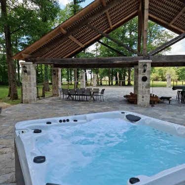 A bubbling hot tub sits on a stone patio near tables and chairs under a large wooden pavilion, surrounded by green trees and an open grassy area.