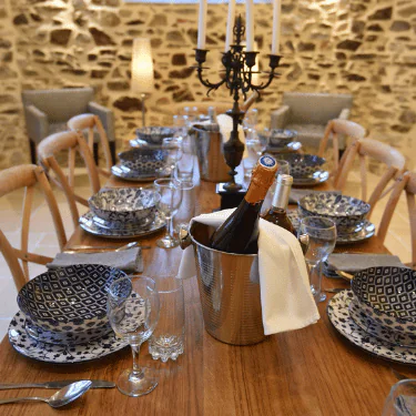 Elegant dining table set for eight with patterned plates, wine glasses, a metal wine cooler holding two bottles, and a black candelabrum; stone wall and armchairs in the background.