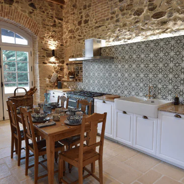 Rustic kitchen with stone walls, patterned tile splashback, white cupboards, wooden dining table set for six, and large window letting in natural light.