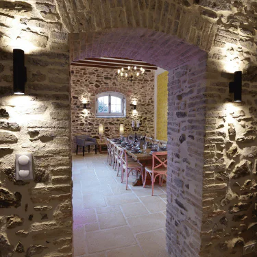 Stone archway leads to a rustic dining room with a long wooden table, pastel chairs, a chandelier, stone walls, and a window letting in natural light. Wall sconces illuminate the textured stone surfaces.
