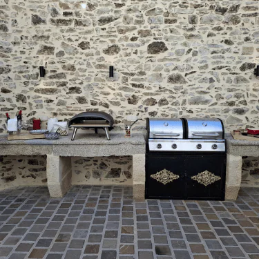 Outdoor stone kitchen with a pizza oven, utensils, and a dual barbecue grill on a stone worktop against a stone wall, with a cobblestone floor in the foreground.