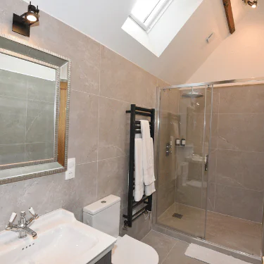 Modern bathroom with grey tiled walls, a walk-in glass shower, white basin, toilet, large mirror, black towel rail with white towels, and a skylight letting in natural light.