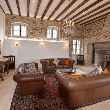 A cosy living room with stone walls, exposed wooden beams, brown leather sofas, a red patterned rug, a chandelier, and large windows letting in natural light.