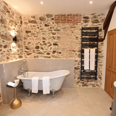 A stylish bathroom with a clawfoot bath, rolled towels, a gold side table, a black towel rail, and stone feature walls, creating a rustic yet elegant atmosphere.