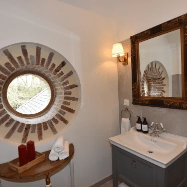A bathroom with a round window framed by bricks, a wooden table with red candles and towels, a washbasin with bottles, a towel ring, a wall lamp, and a large ornate mirror.