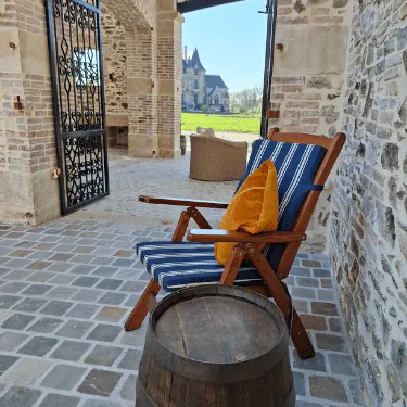 A wooden chair with blue-striped cushions and a yellow pillow sits on a stone patio beside a barrel, with a wrought iron gate and a distant castle visible in the background.
