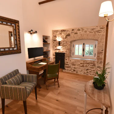 A cosy home office with a wooden desk, computer, and green chair. The room has a striped armchair, wall mirror, stone wall with a window, wooden floor, and a small table with a plant. Warm lighting creates a welcoming feel.