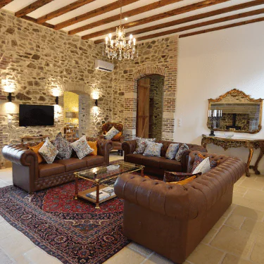 Elegant living room with stone walls, brown leather sofas, a glass coffee table on a red rug, chandelier lighting, a wall-mounted TV, and a decorative console table with a mirror.