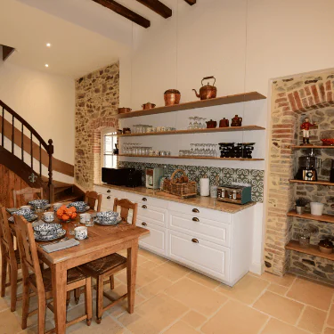 Cosy rustic kitchen with stone walls, wood beams, open shelves holding crockery and copper pots, a wooden dining table set for four, and a staircase visible in the background.