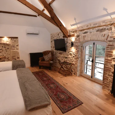 A cosy bedroom with exposed stone walls, wooden beams, two beds, an armchair, a TV, a decorative rug, and a small balcony door letting in natural light.