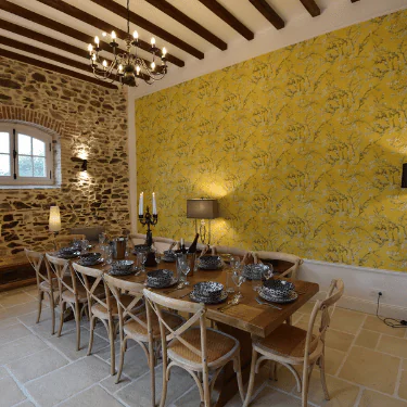 Dining room with a long wooden table set for ten, patterned black and white crockery, stone wall on the left, yellow floral wallpaper on the right, timber beam ceiling, and multiple light fittings.