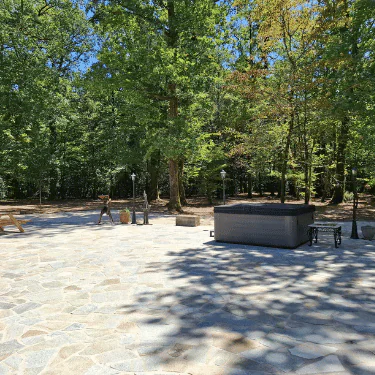 Stone patio with a hot tub, benches, and lamps, surrounded by tall green trees and a forested area under a clear blue sky.