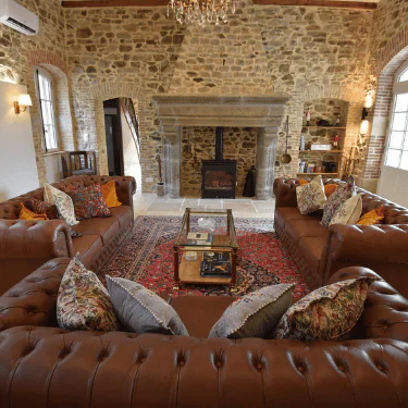 A cosy living room with brown leather sofas, a red patterned rug, a glass coffee table, and a stone fireplace set in a rustic stone wall. Decorative cushions are on the sofas, and shelves line the walls.
